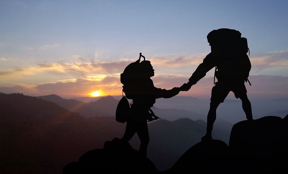 Two hikers' silhouettes on a mountain peak at sunset, one helping the other up.